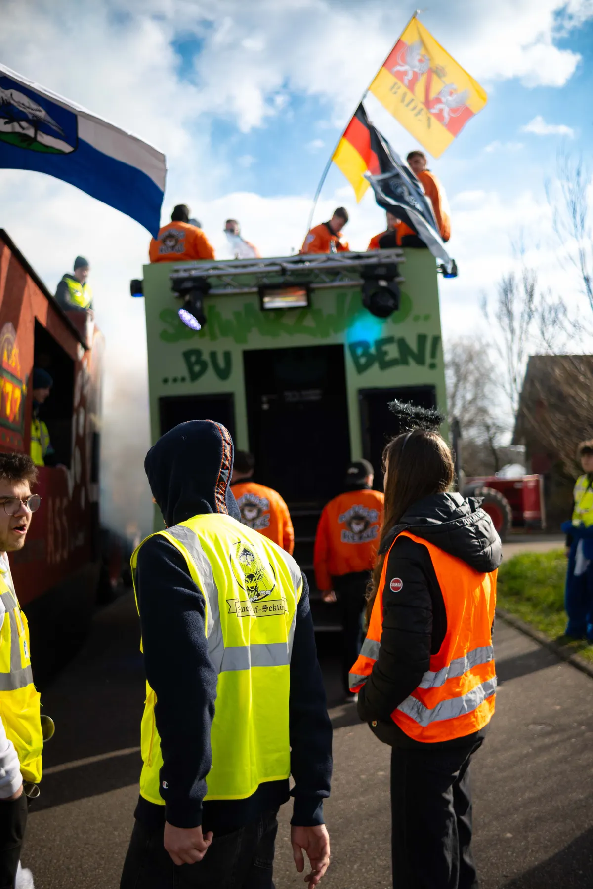 Gruppe von Teilnehmern in Warnwesten auf der Straße, im Hintergrund Vereinsflaggen.