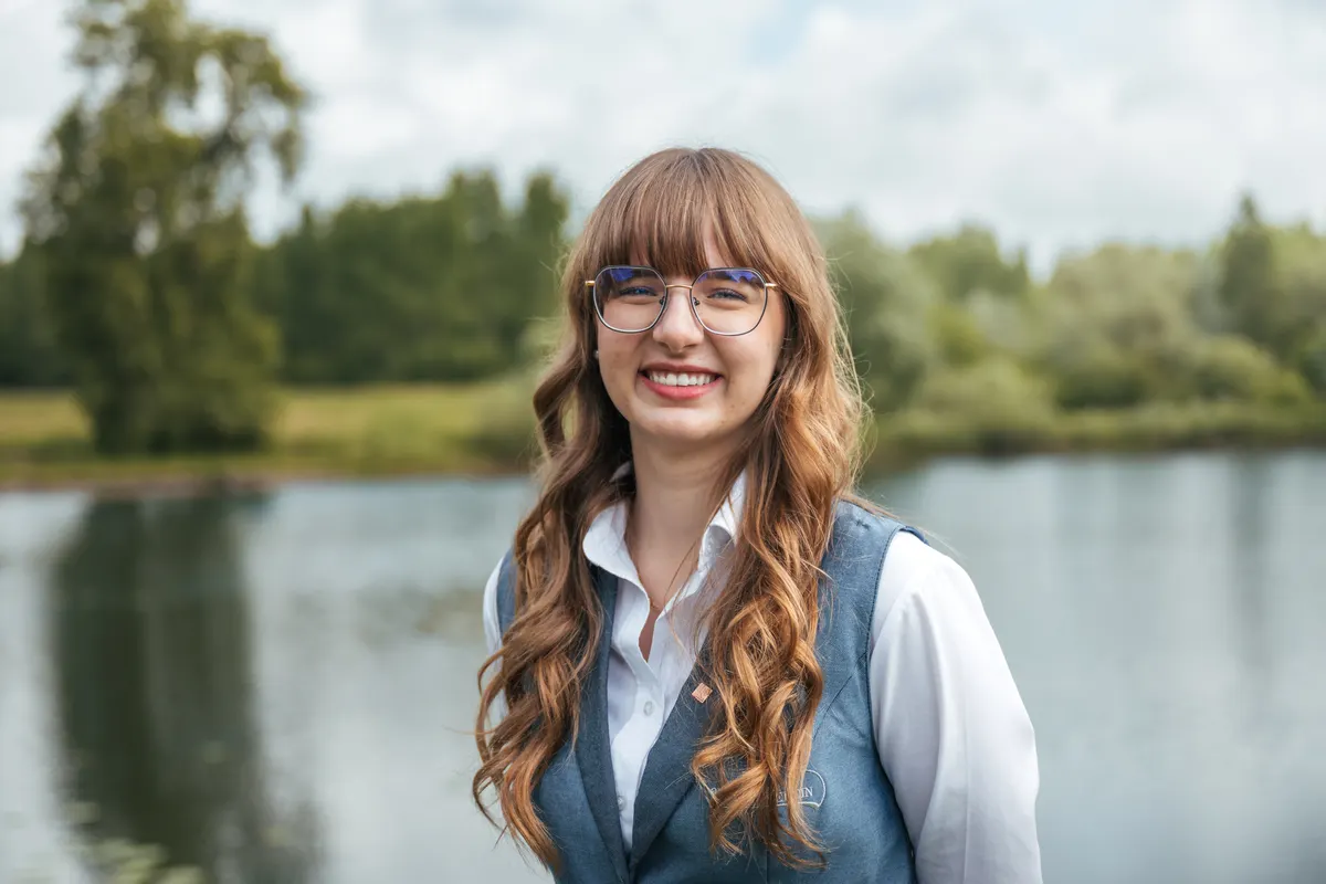 Portrait einer jungen Frau mit langen rotbraunen Haaren und Brille in blau-grauer Trachtenweste am Seeufer.