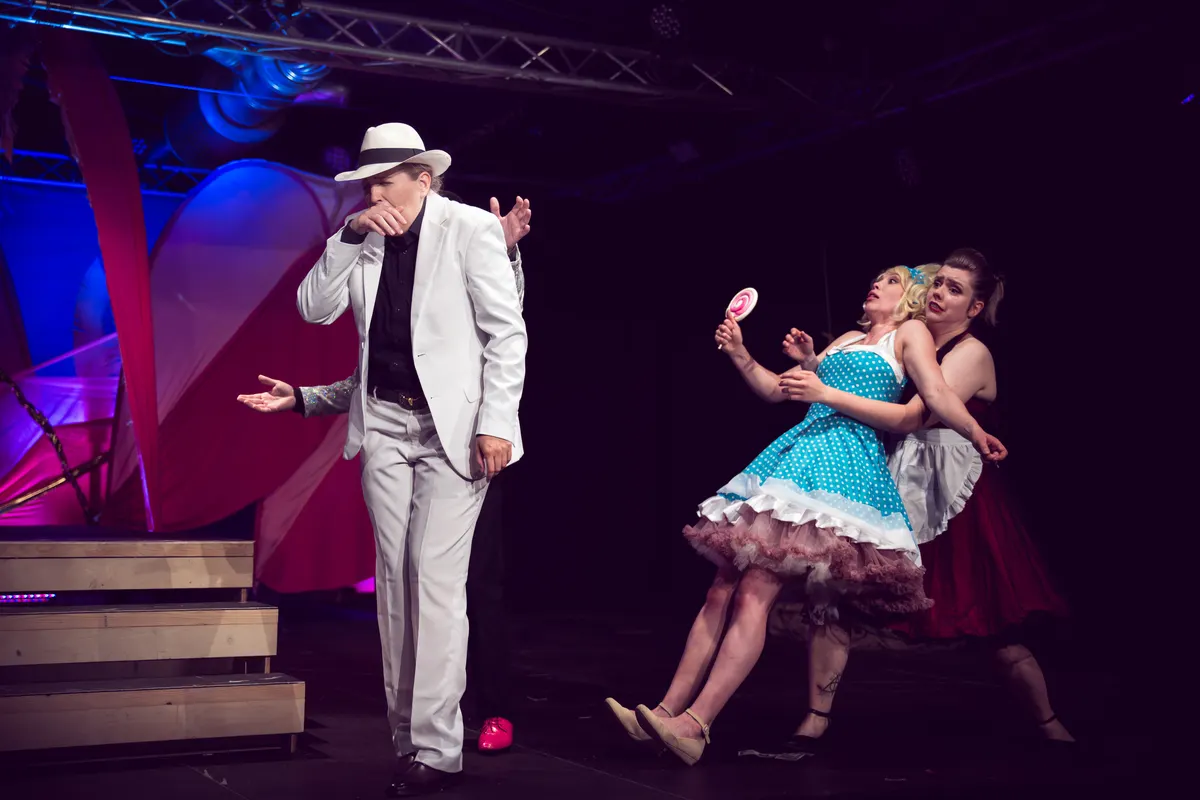 Ein Mann im weißen Anzug tanzt mit zwei Frauen in Petticoat-Kleidern.